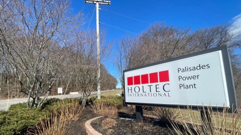 On the right, a white sign with black letters reads Holtec International - Palisades Power Plant, while a road on the left leads up the hill past trees with no leaves. The entrance to the now shuttered nuclear plant in Covert Township, Michigan.