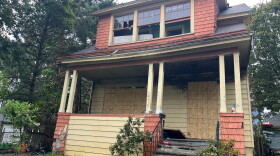 This is a photo of the red and orange house where the fire broke out. Two of the the three front-facing windows on the second floor are broken. The third, on the right, was left open. There's some charred pieces in the driveway. The door and front floor windows are boarded up. You can see fire damage on the exterior wall above the door. 