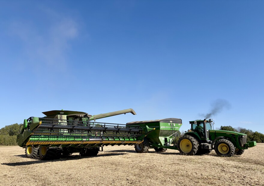 A green John Deere combine unloads soybeans into the wagon hitched to a green tractor. 