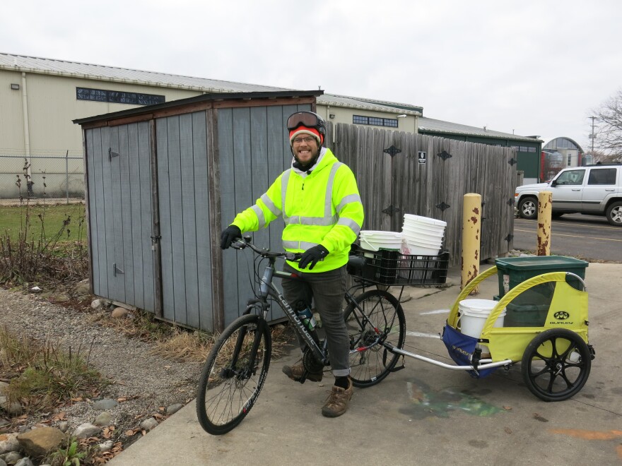 A man in a bright yellow jacket stands with a bike, which has a trailer loaded with bins and buckets.