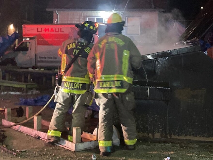 Two Ann Arbor firefighters stand in front of a smoldering dumpster, using a firehose to douse a fire inside the dumpster. 