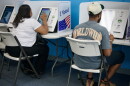 Interior photo that shows two people sitting in folding chairs with their backs to the camera. They appear to be making selections on electronic screens that are voting machines.