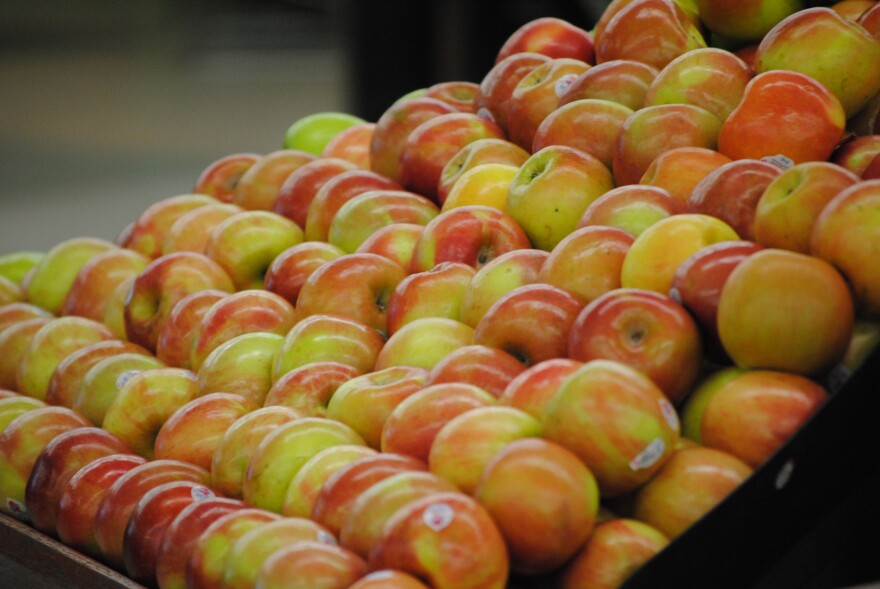 Several dozen green-reddish apples are stacked next to each other in a produce bin. 