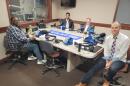 Four men sit at a table in a radio talk studio: a man front left has short dark hair and a dark beard and is wearing glasses, a blue plaid button-down shirt, jeans and sneakers; a man front right has short dark hair and is wearing a white button-down shirt, pink and blue plaid tie, jeans and brown shoes; a man back left has short dark hair and a dark beard and is wearing a grey jackets over a light blue button-down shirt; a man back right has short grey hair and is wearing a grey blazer, white button-down shirt and grey tie.