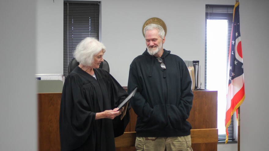 A woman in judges robes holds a certificate as she turns toward a man in a hoodie. They stand at the front of a small courtroom, with an Ohio flag to one side. 