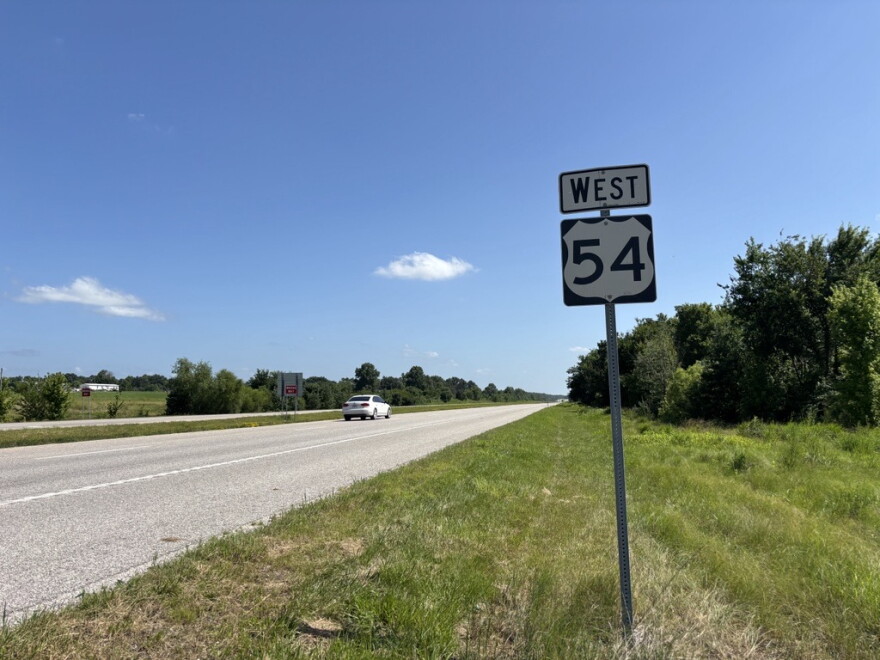 A wide shot of Highway 54 in rural Mid-Missouri. A "West 54" road sign stand in the ditch. The grass on the side of the road is green. A white sedan travels along the highway in the distance. 