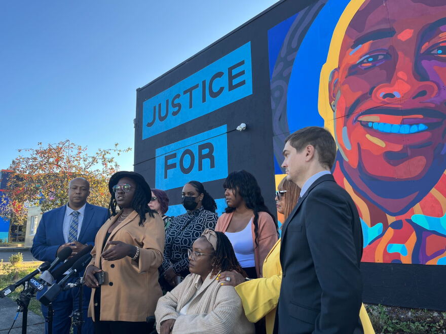 A group of people stand, with one person sitting, in front of several microphones with a colorful mural of a man's face and the words "Justice For" appearing behind them.