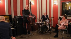 A pianist, bass player and drummer in a red music studio talk with a woman sitting nearby in a pink dress.