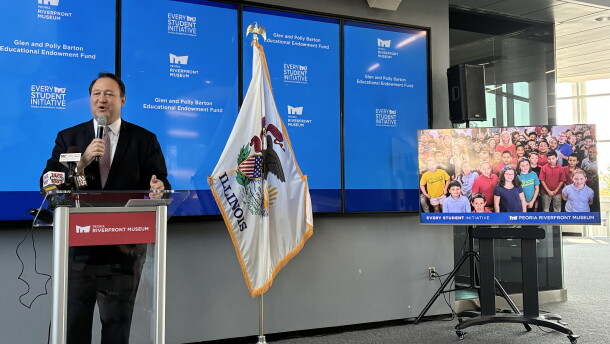 A man speaks at a podium beside an Illinois flag, with multiple screens behind him displaying “Every Student Initiative.” A monitor shows a group photo of children.