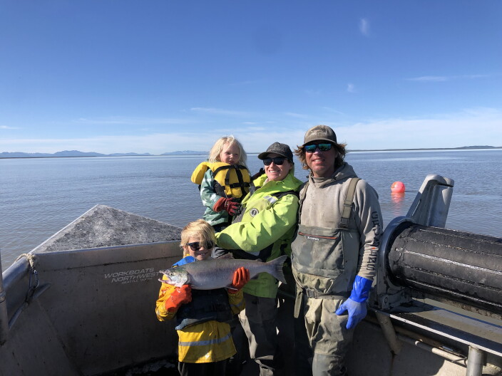 A family, two adults and two young blonde kids, pose on a fishing boat with blue bay water behind them.
