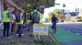 A bicyclist rides in a green bike lane along Prince Avenue while several people in safety vests stand beside a sign requesting feedback on a pilot project.