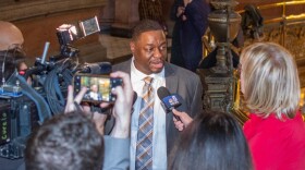 A man in a suit and tie, identified as Sen. Emil Jones III, addresses a swarm of eager reporters with microphones and cameras in a hallway. As the staircase with an ornate railing looms behind him, he discusses the ongoing federal corruption probe related to red-light cameras.