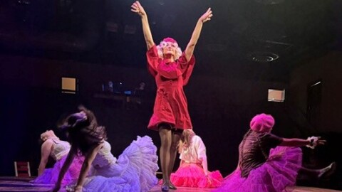 A woman wearing red with a blond wig standing above a circle of women in dance outfits.