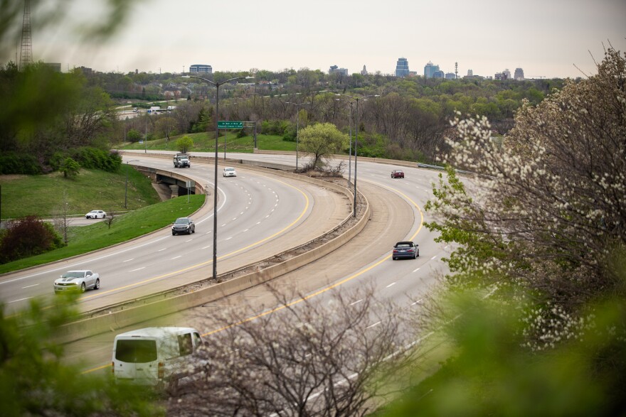 A curving highway is shown surrounded by trees and grassy areas. Another smaller road can be seen at far left cutting below the curing highway. In far background, the Kansas City skyline can be seen.