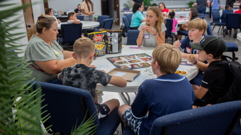 A group of five kids is sitting at a circular folding table with an adult eating chips and popcorn. They're playing the classic board game Clue.