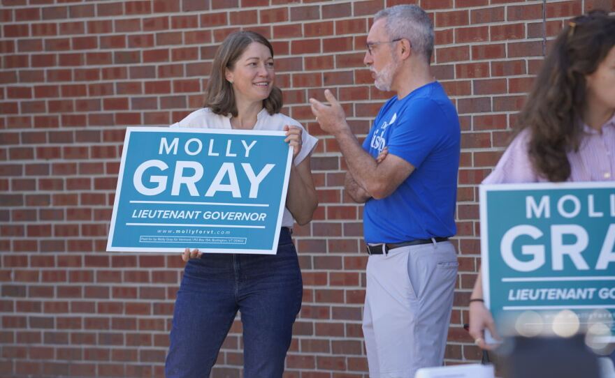 A woman holds a sign reading "Molly Gray, lieutenant governor" and talks with another person