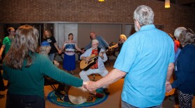 A group of dances stand in a circle holding hands, surrounding a seated guitar player in the center of the circle.