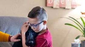 A young boy, wearing blue-rimmed rectangular glasses, cradles the head of his black lab dog. The boy wears a red t-shirt. His mother's arm can be seen patting the dog, wearing a yellow sweater. They sit on a grey couch against a backdrop of a tan wall with an out-of-focus wall hanging and a plant and tissue box visible.