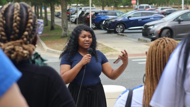 Jasmine Brown speaks into a microphone while addressing a small crowd during a downtown Pensacola rally in May 2025. She holds a phone in one hand and gestures as she talks, with parked cars and trees visible in the background.