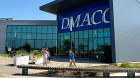 A woman wearing a backpack walks out of a large building with the words "DMACC" and "student center." She is accompanied by a toddler girl and an older boy. Two other figures can be seen in the background. People leave the Student Center building on Monday, July 28, 2025, at the Ankeny campus of Des Moines Area Community College in Ankeny, Iowa.