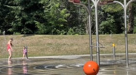  A woman and her 4-year-old daughter stand in bathing suits near sprinklers, getting wet in a concrete basin on a sunny day at Northacres Park in Seattle. 