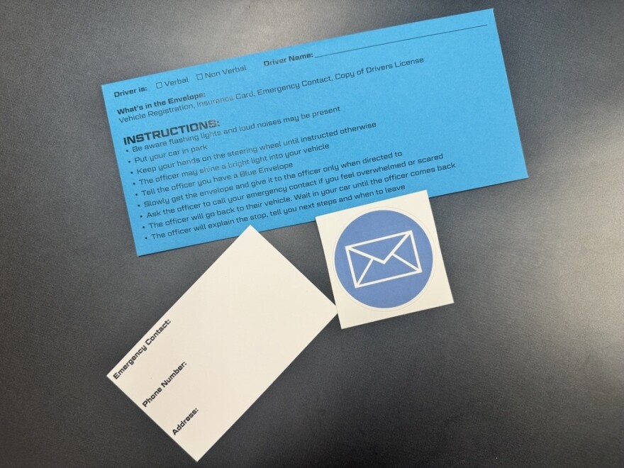 A blue envelope for autistic drivers with instructions on the front, splayed out on a table with an emergency contact card and a blue envelope sticker.
