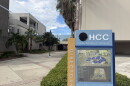 A blue and tan directory sign with a map of Hillsborough College's Dale Mabry campus stands in front of the entrance of grey classroom buildings and trees lining the walkway.