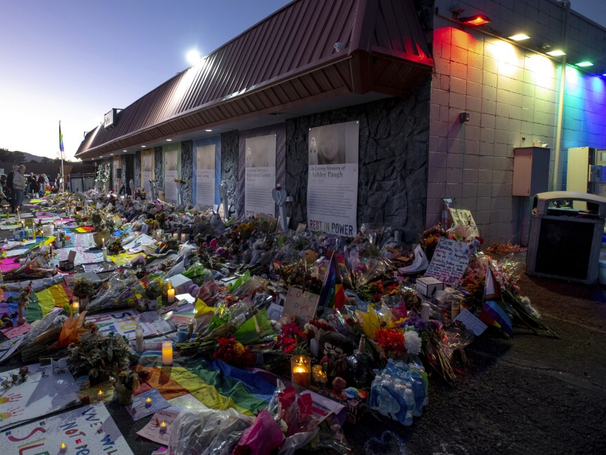 A large memorial, full of colorful flowers and signs, reaches up to the doorway in front of Club Q, site of a mass shooting. The building is viewed from a side angle.