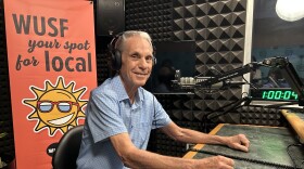 Older man sits in front of microphone with book on studio desk with WUSF Your Spot for Local sign. 