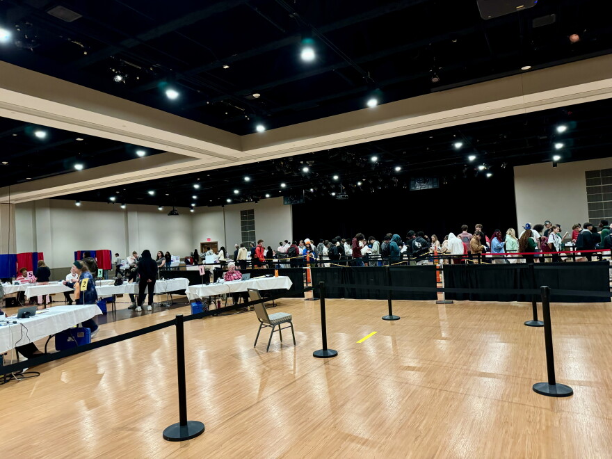 A large room with people lined up at tables, possibly for registration or a similar event. The space is well-lit with overhead lights, and several barriers guide the flow of the queue. Tables with chairs are scattered throughout.