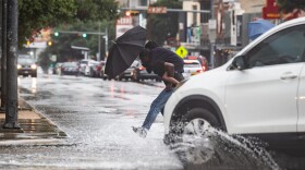 A man carries another person on his back with an umbrella across a flooded sidewalk.