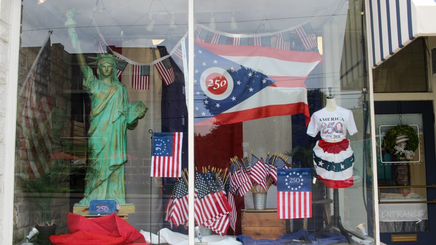 A storefront has a mini Statue of Liberty and many American flags in the window, including several flags that say "250" or '76. The largest flag is an Ohio flag with "250" in the center of the circle. A mannequin wears an America 250 shirt and a skirt made out of red, white, and blue bunting. 