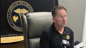 Peoria County Coroner Jamie Harwood speaks into media microphones with a banner behind his back at a 2024 news conference in his office.