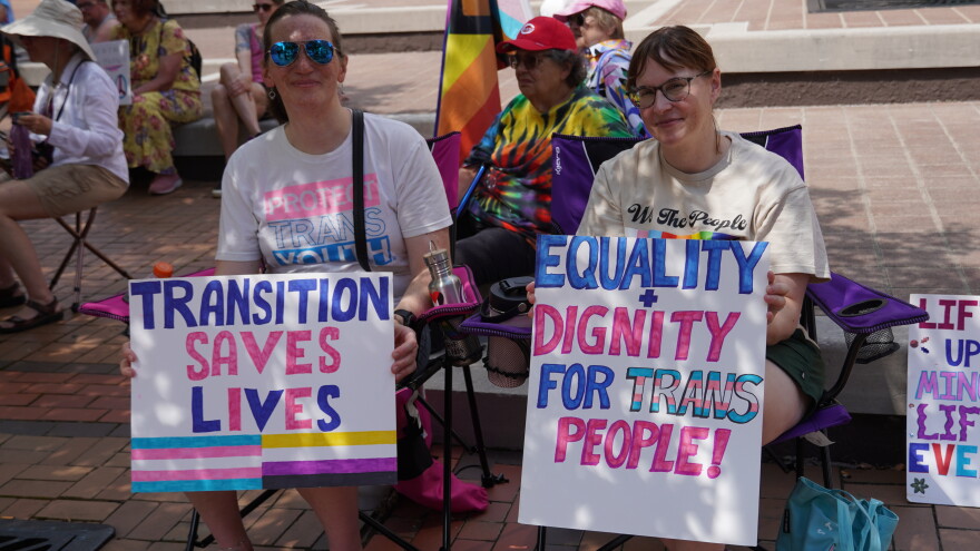 image of 2 people holding signs that say "Transition saves lives" and "Equality plus dignity for Trans People"