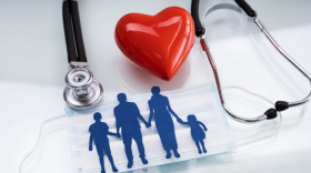 Un recorte de una familia tomada de la mano se encuentra encima de una mascarilla con un estetoscopio y un corazón en el fondo.