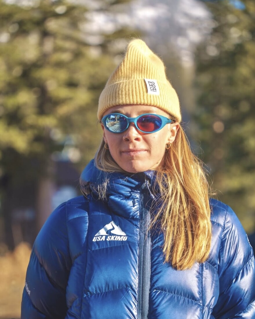 A young woman in sunglasses and a beanie stands in a big puffy coat with a USA Skimo logo.