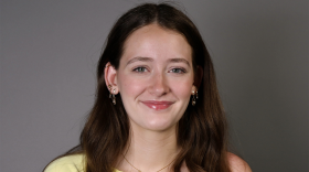 A young woman with long brown hair wears a yellow shirt and smiles in front of a grey background