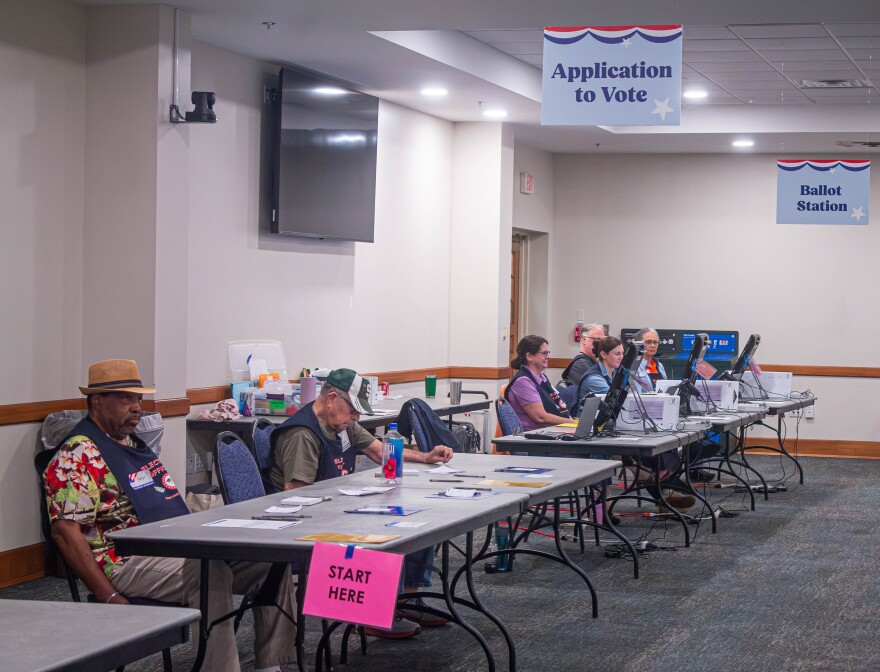 Six election workers are seated at two folding tables labeled "Application to Vote" and "Ballot Station." Nobody is in line to vote early.