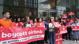 District 1 Supervisor Connie Chan speaks at a protest in front of Airbnb wearing a white shirt and jeans, surrounded by red and white protests signs that read "boycott airbnb."