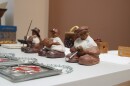 Three clay sculptures of fat men holding different sports equipment sit on a table. In front of them are stained glass red flowers. Behind them are wooden blocks.