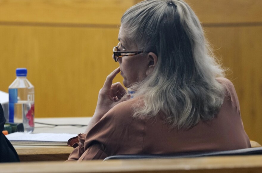 A woman with draying hair sits calmly in a courtroom as testimony occurs. She sits with one finger resting on her upper lip.