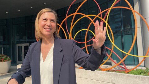 U.S. Senator Elissa Slotkin stands and smiles over her left shoulder as she waves. She's standing in front of a scultpure with yellow, orange and red circles. Behind that is a door to enter the Helen Devos Children's Hospital in Grand Rapids.