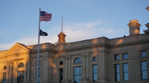 The sun sets on a sandstone building with the U.S. and a P.O.W.//M.I.A flag in front of it.