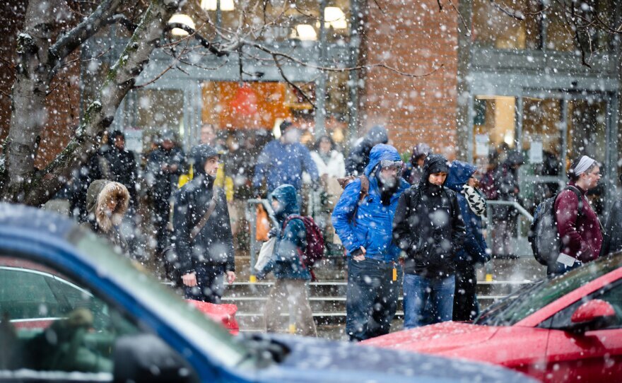 Snow falls as cars and people move across a street.