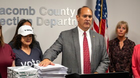 Macomb County Clerk and Republican candidate for Michigan secretary of state Anthony Forlini speaks at a podium in a county elections office. He stands before stacks of documents and boxes of copy paper, flanked by several people. American and Michigan flags stand behind him, with election-office signage on the wall.