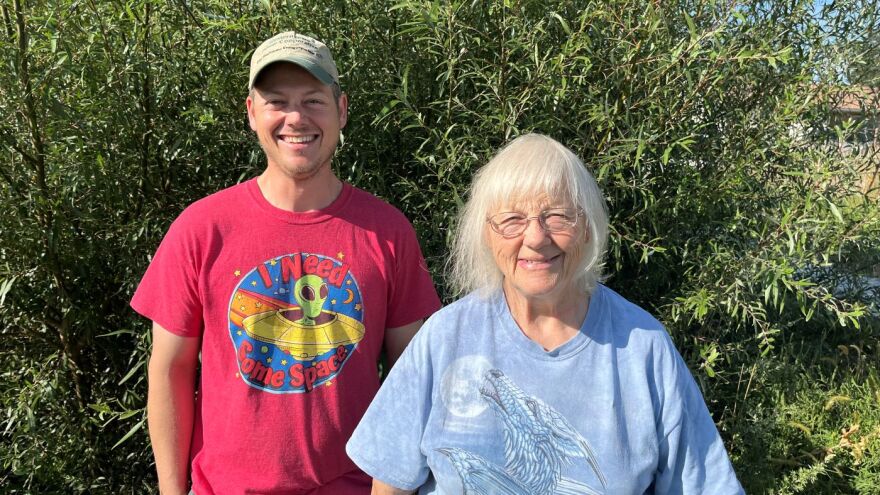 There is an older day to the right wearing a light blue-ish shirt with a dragon-wolf howling at a moon. She has grey hair and classes. A younger man is to the left. He is wearing a t-shirt with an alien on it that says "I Need Some Space." He is smiling with a lighter green baseball cap. He is smiling too.