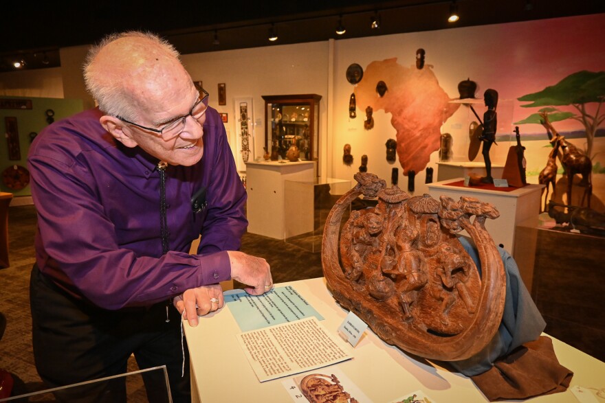 A man wearing a purple shirt leans on a display box looking at a wooden carving. Behind him are many different African artifacts and sculptures.