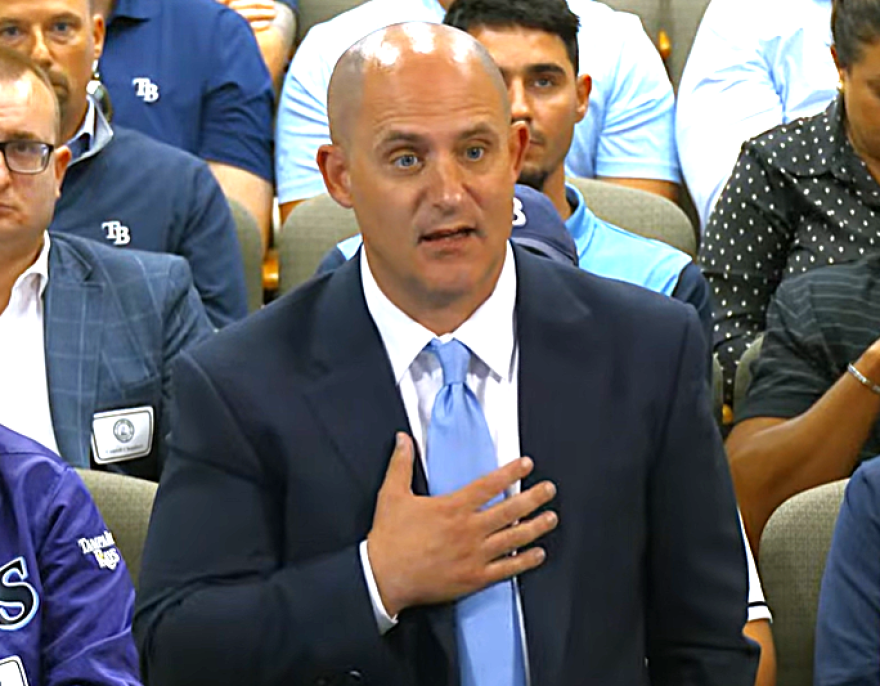 bald white man in blue suit sand light blue tie peaks with his right hand across his chest 