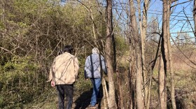 Photo of two people walking away from the camera. They are outdoors in a wooded area. It's very early spring, so there is more brown than green, though some small new leaves are visible on some tree branches.