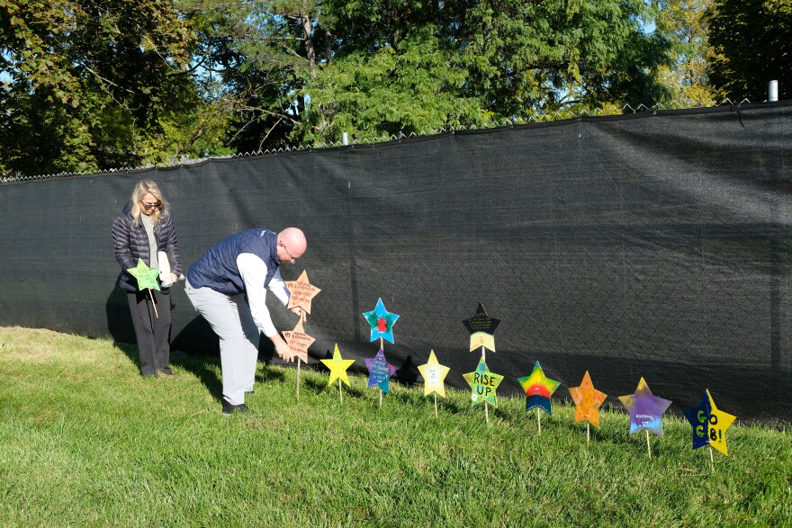 A man in a navy vest and button down shirt pushes a stake with two stars on it into the grassy ground, while a woman in a black jacket stands behind him holding another star.